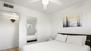 Un dormitorio con una cama grande, una mesita de noche, un espejo, un ventilador de techo y una obra de arte colgada en la pared.