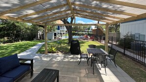 A covered patio area with a wooden pergola, outdoor seating, and a view of a backyard with a pool and a house. - **PRIVATE POOL** 1 Block to Beach (Kure Beach)