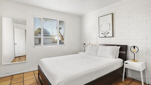 Un dormitorio con cama, mesita de noche, un cuadro en la pared y una ventana con persianas.