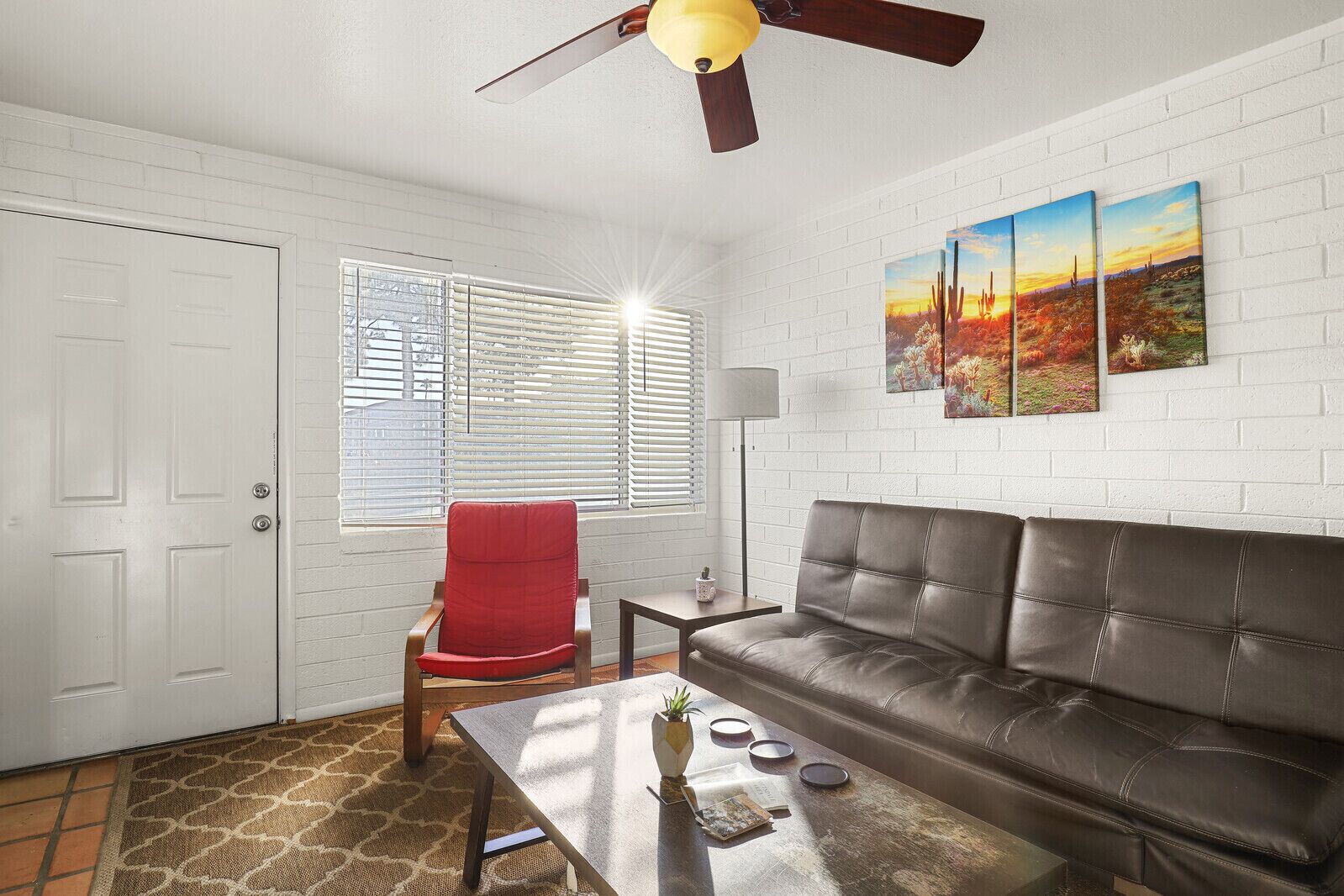 A living room with a brown leather sofa, a red chair, a coffee table, and a ceiling fan. There are three paintings on the wall and a door with a window.
