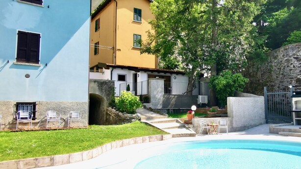 A swimming pool with a stone border, a grassy area, and a building with a yellow facade and a stone wall. - BLUE HOUSE 3 by Design Studio (Bellano)