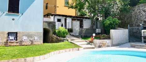 Une piscine bordée de pierres, une zone herbeuse et un bâtiment à la façade jaune et au mur en pierre.