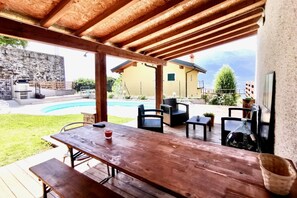 Un patio al aire libre con mesas y sillas de madera, una piscina y un muro de piedra al fondo.