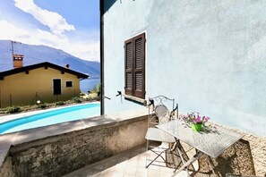 Ein Poolbereich mit Tisch und Stühlen, ein Fenster mit Rollläden und im Hintergrund eine Bergblick und ein Gebäude.