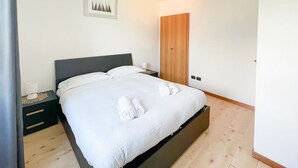 A neatly made bed with white linens and rolled towels, a bedside table with a lamp, and a wooden wardrobe in a minimalistic room. - BLUE HOUSE 3 by Design Studio (Bellano)