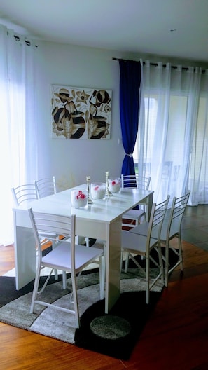 Uma sala de jantar com uma mesa e cadeiras brancas, um quadro na parede e uma cortina azul.