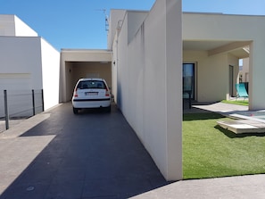 Um edifício residencial moderno com um carro branco estacionado na entrada, uma pequena área de pátio com gramado verde e um céu azul claro.