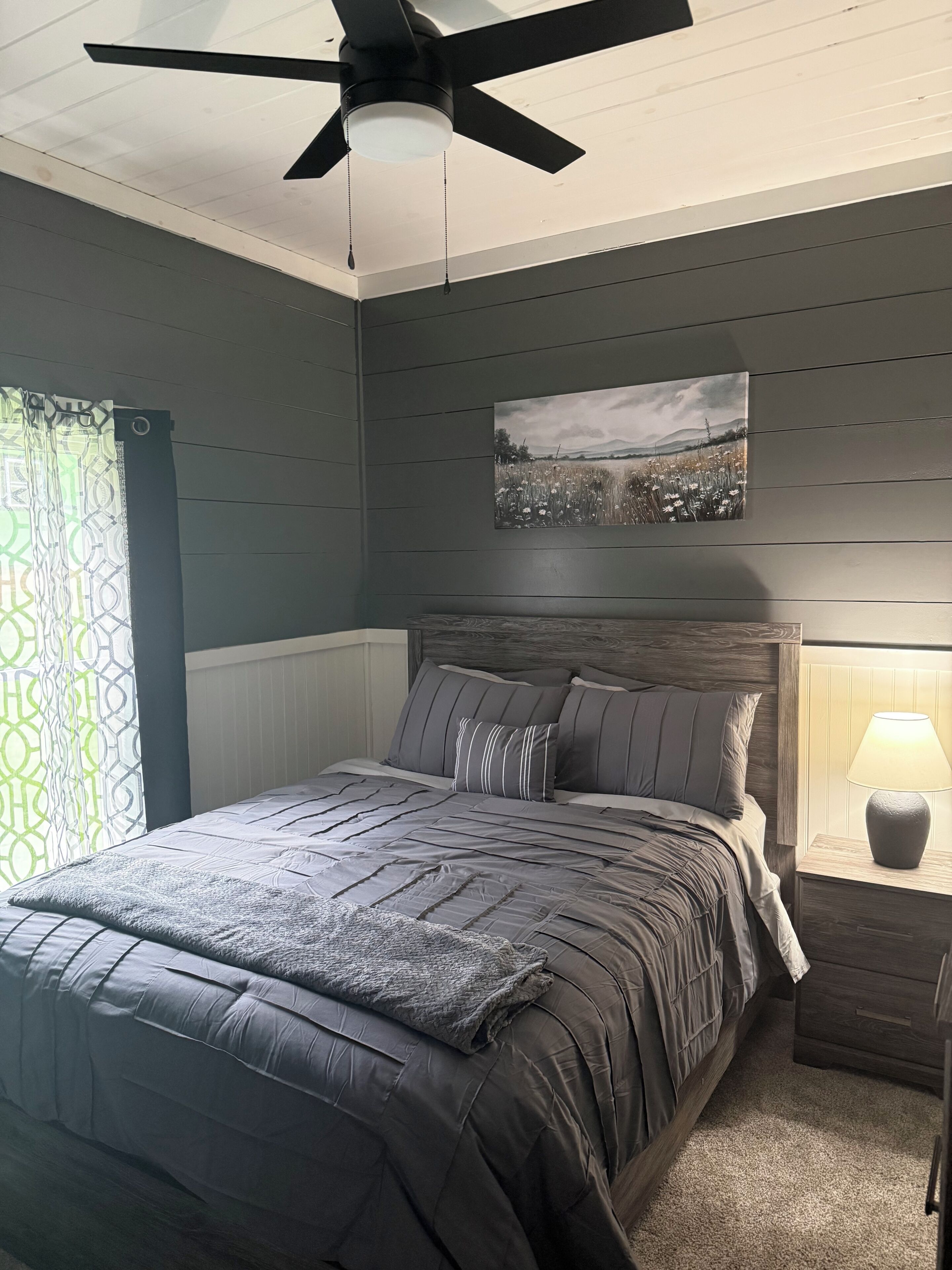 Un dormitorio con cama, ventilador de techo, mesita de noche con lámpara y un cuadro en la pared.
