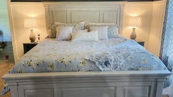Un dormitorio con una cama grande, dos mesitas de noche con lámparas, un adorno en forma de corazón en la pared y un estante pequeño con fotografías.