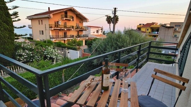 A balcony with a wooden table and chairs, overlooking a residential area with houses and greenery. - Villetta Gina (Avola)