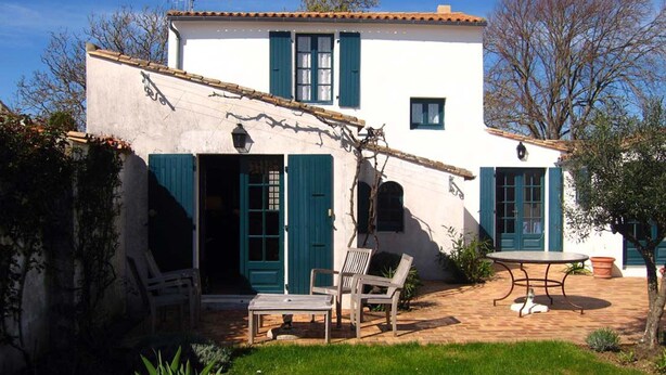 Ein weißes Gebäude mit grünen Fensterläden, eine Terrasse mit Tisch und Stühlen und ein Garten mit Pflanzen.