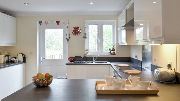 A modern kitchen with white cabinets, a central island with a tray of white cups, a kettle, and a bowl of fruit. - 3 bedroom accommodation in Bampton, near Tiverton (Bampton, near Tiverton)