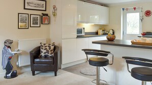 A modern kitchen with white cabinets, a microwave, and a kettle. There are two bar stools and a brown leather sofa with patterned cushions. - 3 bedroom accommodation in Bampton, near Tiverton (Bampton, near Tiverton)