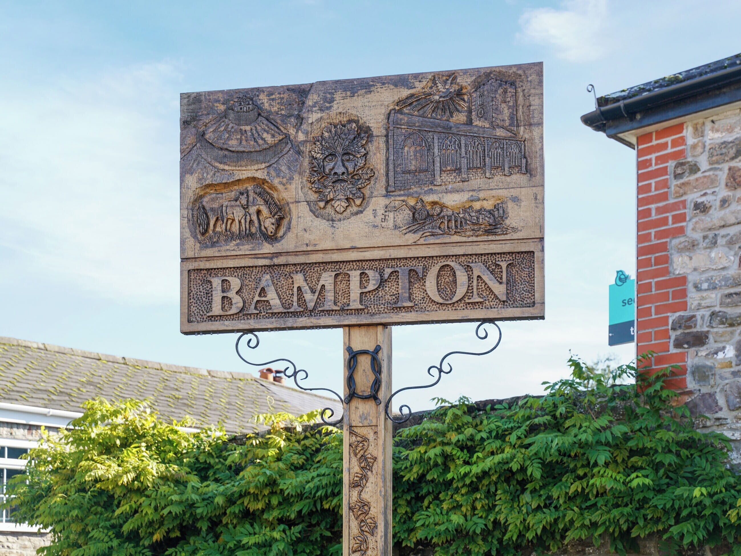 Een houten bord met 'BAMPTON' en diverse houtsnijwerken, waaronder een leeuw en gebouwen, bevestigd aan een paal met een sierlijk ijzeren ontwerp.