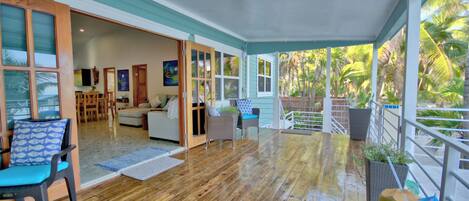 A covered porch with wooden flooring, a dining area with chairs and a table, and a view of palm trees.