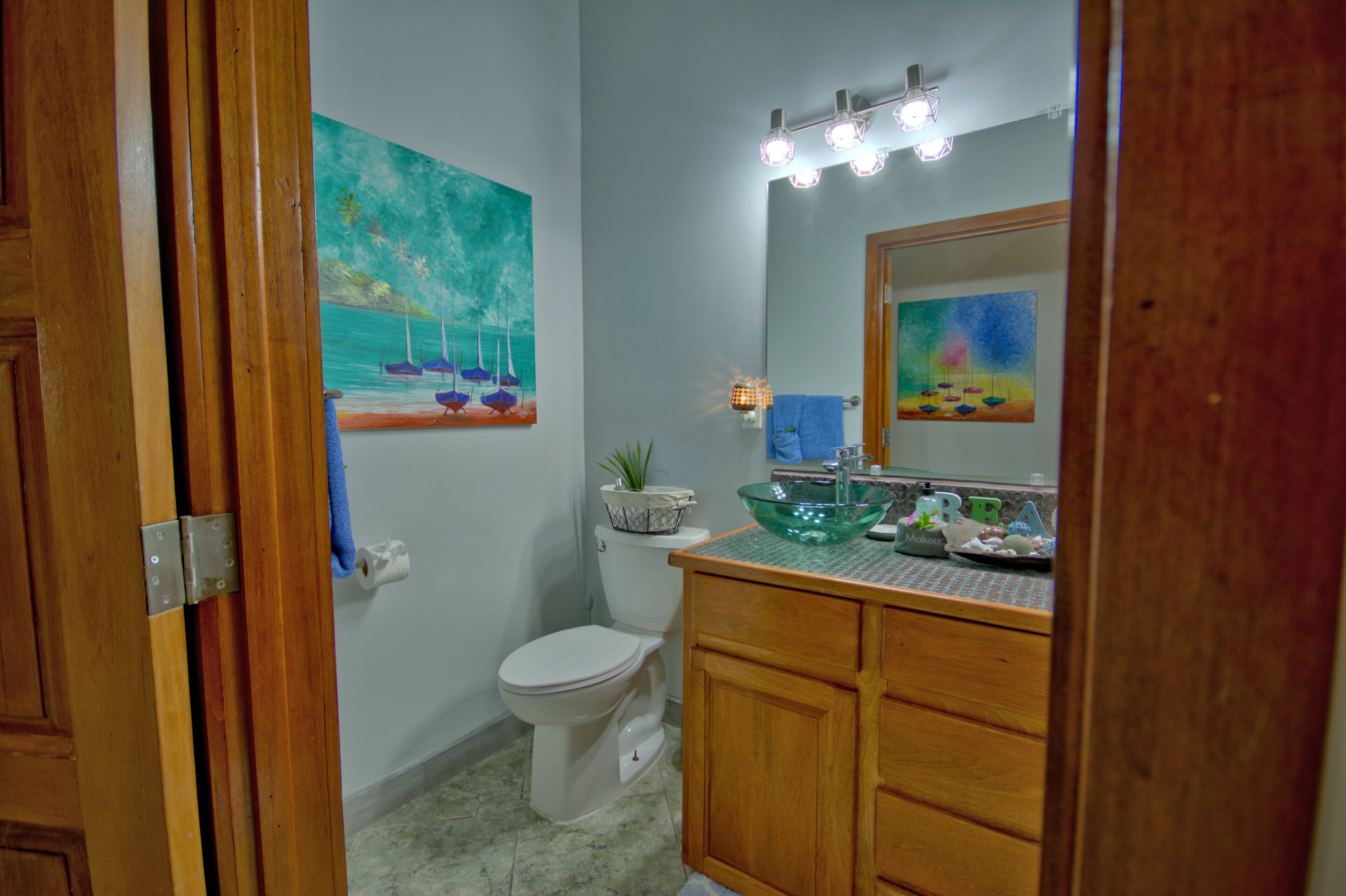 A bathroom with a wooden vanity, a glass bowl sink, a toilet, and two paintings on the walls.
