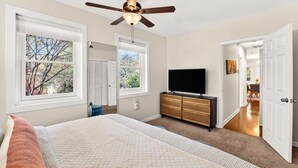 A bedroom with a bed, a ceiling fan, a TV on a wooden cabinet, and a view of the outside through a window. - Stylish Inman Park Condo, Walk to Beltline (Atlanta)
