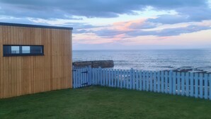 A wooden building with a small window, a grassy lawn, and a white fence overlooking a body of water. - 1 bedroom accommodation in Hilton, near Tain (Hilton, near Tain)