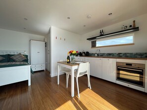 Une cuisine moderne avec un coin repas, un plancher en bois, des meubles blancs et une fenêtre au-dessus de l’évier.