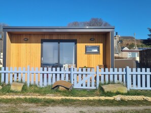 Une petite cabane en bois avec une porte coulissante en verre, une petite fenêtre et une clôture blanche à piquets devant.