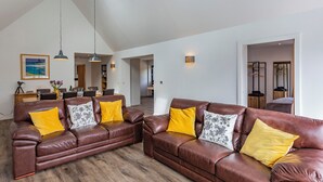 Ein geräumiges Wohnzimmer mit zwei braunen Ledersofas, gelben und gemusterten Kissen, einem Essbereich mit Tisch und Stühlen sowie einer Küche im Hintergrund.