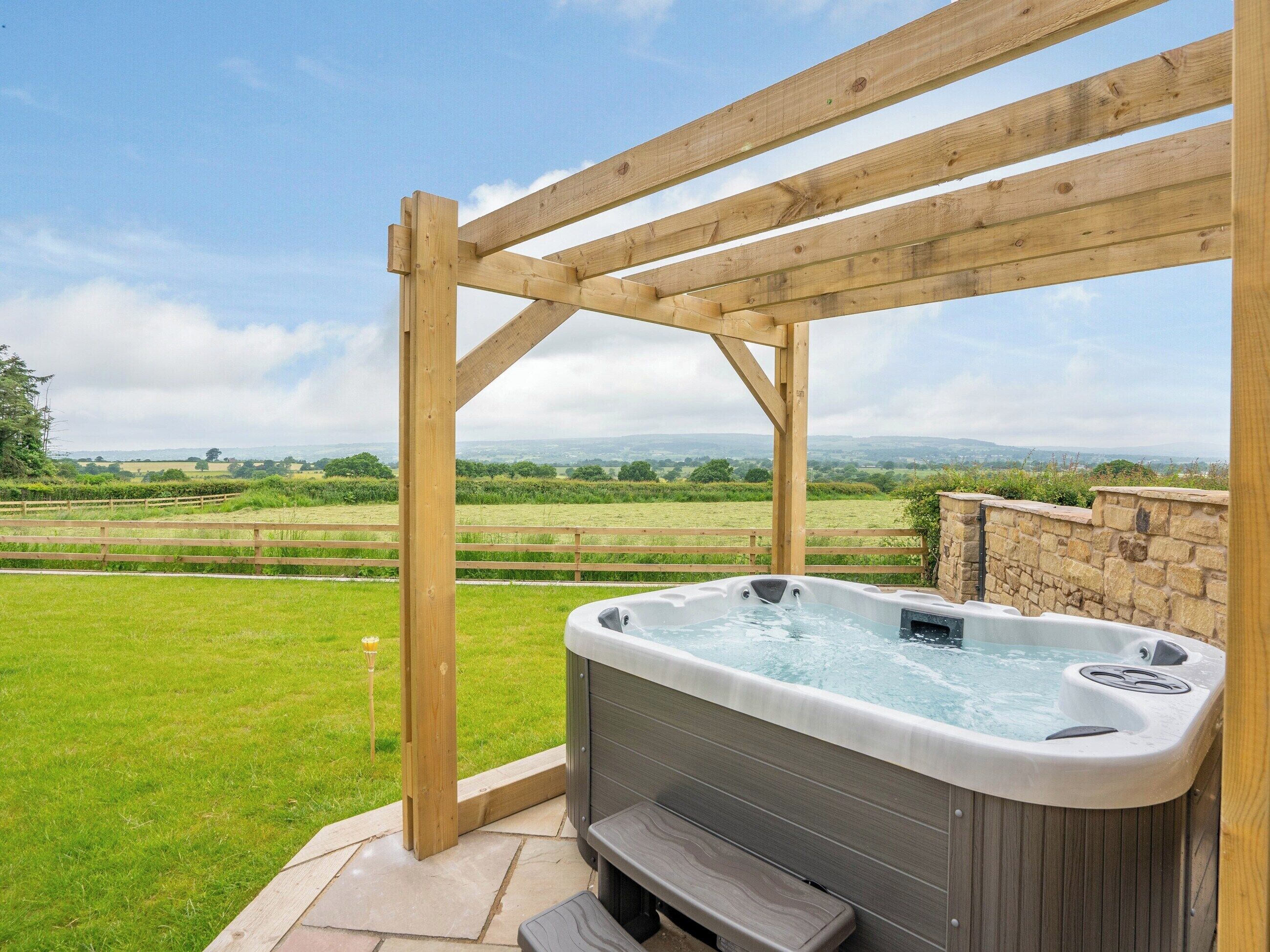 Un bain à remous installé sous une pergola en bois, offrant une vue sur un champ verdoyant et des collines lointaines.
