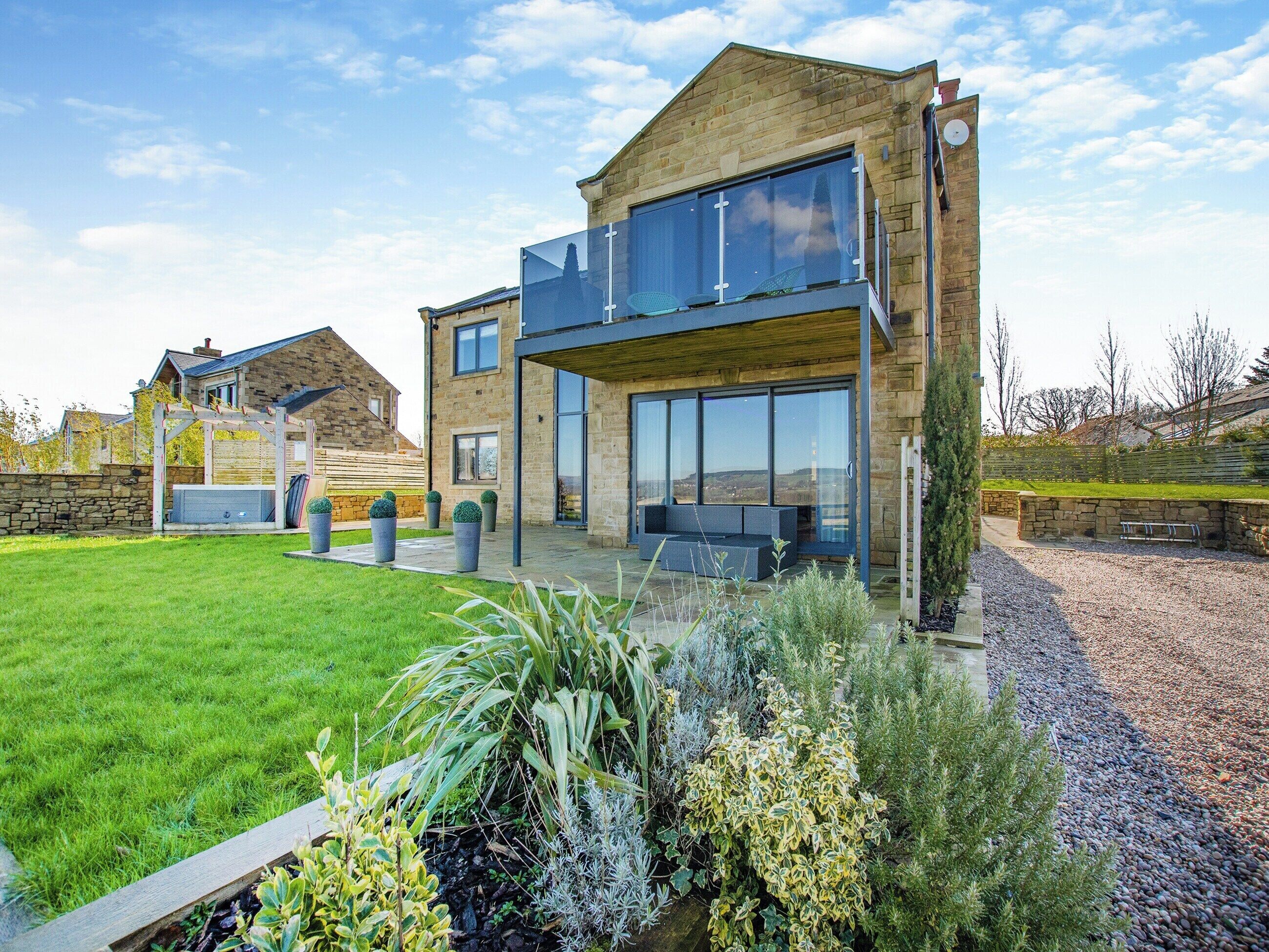 Une maison moderne à deux étages, dotée d’un grand balcon en verre, d’une façade en pierre et d’une pelouse bien entretenue.