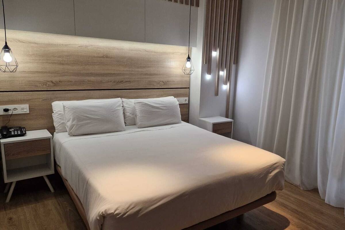Quarto de hotel moderno com cabeceira de madeira, uma cama grande com roupa de cama branca, duas mesas de cabeceira e um criado-mudo com telefone e carregador.