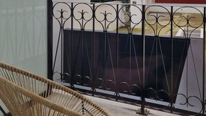 Un balcón con una silla de mimbre, una barandilla de metal y vistas a edificios y follaje.