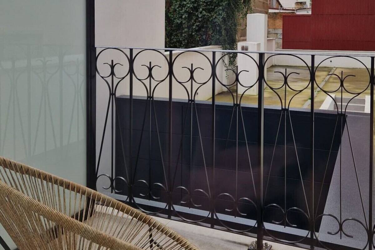 Varanda com cadeira de vime, grade de metal e vista para prédios e vegetação.