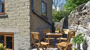 A stone building with a wooden table and chairs, a bottle of champagne, and flowers. - The Old Candle House (Longnor, near Buxton)