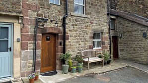 A stone building with a wooden door, a bench, and potted plants on the front. - The Old Candle House (Longnor, near Buxton)