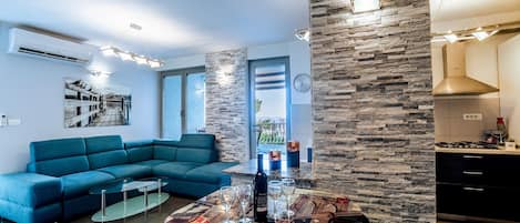 Een moderne woonkamer met een blauwe bank, een ronde tafel met een tafelkleed met New York-thema, en een keukenhoek met een stenen muur en een ingebouwde kookplaat.