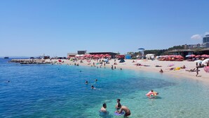 A beach with clear blue water, people swimming and sunbathing, and red umbrellas. - Modern apt Close to Znjan Beach, With big Terrace (Split)