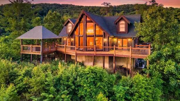 A log cabin with a large deck and a covered area, surrounded by dense forest. - 🍁SPECIAL⛄️: LUX Log Mansion w/180° VIEWS, SAUNA, & HOT TUB! 30min to Dollywood, (Sevierville)