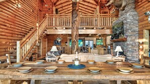A spacious log cabin interior with a dining area set for a meal, a fireplace, and a staircase leading to a balcony. - 🍁SPECIAL⛄️: LUX Log Mansion w/180° VIEWS, SAUNA, & HOT TUB! 30min to Dollywood, (Sevierville)