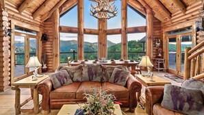 A log cabin interior with a large window, a chandelier, a leather sofa, and a dining table. - 🍁SPECIAL⛄️: LUX Log Mansion w/180° VIEWS, SAUNA, & HOT TUB! 30min to Dollywood, (Sevierville)