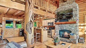 A log cabin interior with a stone fireplace, wooden furniture, and a kitchen area. - 🍁SPECIAL⛄️: LUX Log Mansion w/180° VIEWS, SAUNA, & HOT TUB! 30min to Dollywood, (Sevierville)