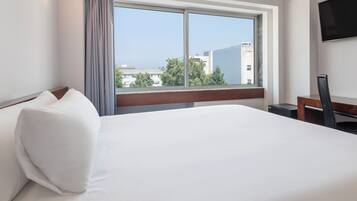 A hotel room with a large bed, a desk, a chair, a TV, and a window with a city view.
