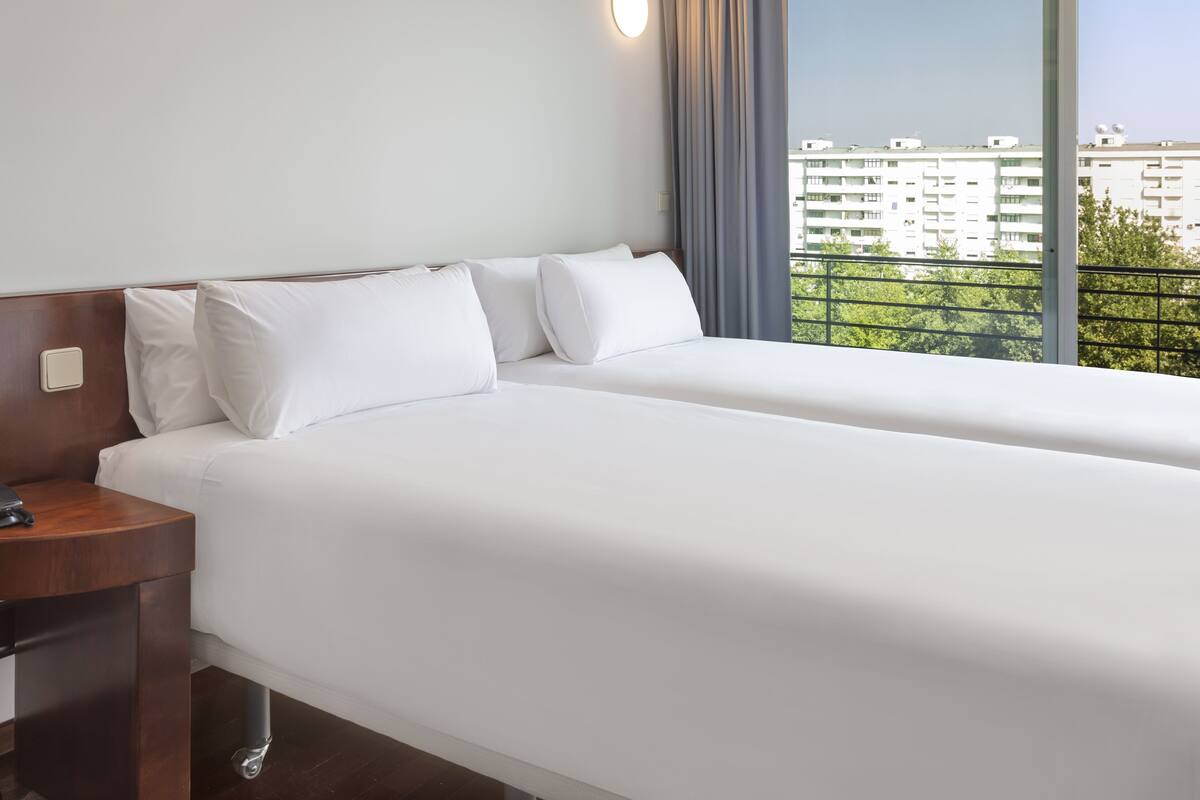 Een hotelkamer met twee eenpersoonsbedden, een houten nachtkastje, een wandlamp en uitzicht op een stadsbeeld via een balkon.