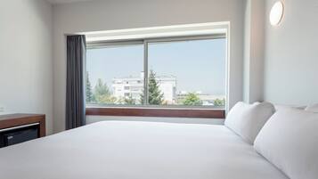 A hotel room with a large bed, a window with curtains, a bedside table, and a wall-mounted light.