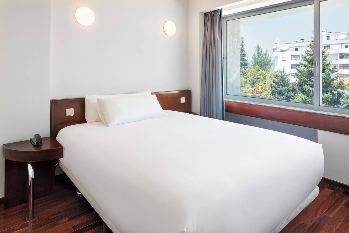 Een hotelkamer met een groot bed, een nachtkastje met een telefoon, en een raam met uitzicht op gebouwen en bomen.