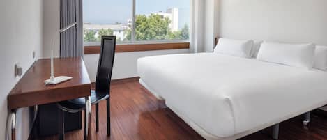A hotel room with a large bed, a desk with a chair, a window with a view of buildings and trees, and a flat-screen TV mounted on the wall.