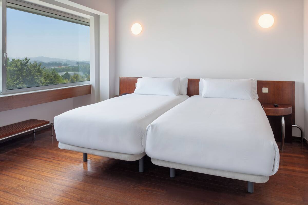 Een hotelkamer met twee eenpersoonsbedden, een houten bankje, een nachtkastje en een groot raam met uitzicht op groen en heuvels.