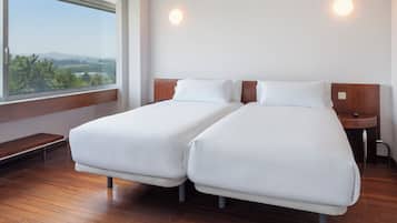 A hotel room with two single beds, a wooden bench, a nightstand, and a large window with a view of greenery and hills.