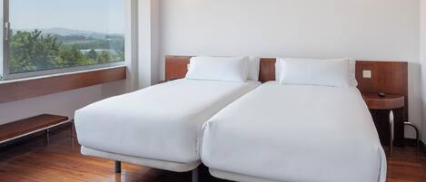 Habitación de hotel con dos camas individuales, un banco de madera, una mesita de noche y un ventanal con vistas a zonas verdes y colinas.