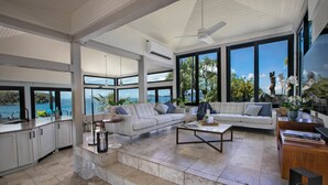 Amplia sala de estar con un gran ventanal con vista al mar, un sofá blanco, una mesa de centro y una zona de cocina con armarios blancos.