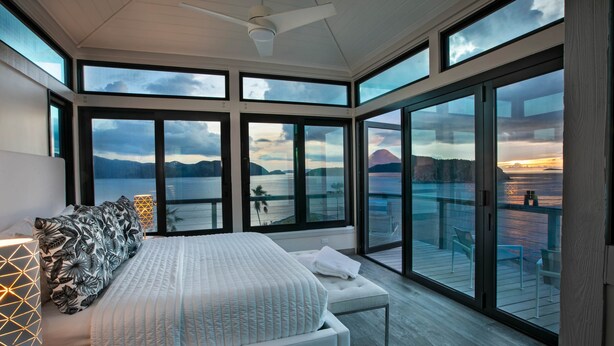 Un dormitorio moderno con una cama grande, una mesita de noche con lámpara y vistas al mar a través de amplios ventanales.