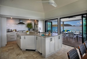 Moderni keittiö, jossa on suuri keittiösaareke, valkoiset kaapit ja suuret liukuovet, joista avautuu näkymä merelle.