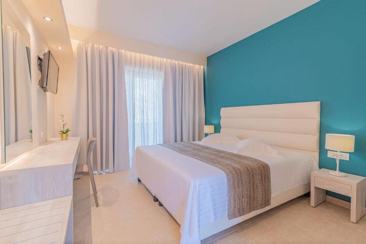 Un dormitorio moderno con una cama grande, una mesita de noche con lámpara, un escritorio con silla y un amplio ventanal con cortinas.